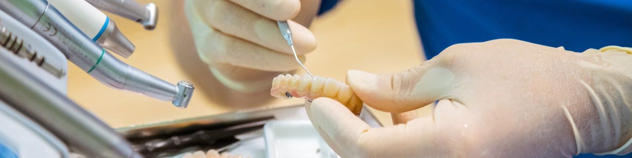 Nahaufnahme von Händen in Handschuhen, die mit einem zahnärztlichen Instrument an einer Zahnprothese arbeiten.