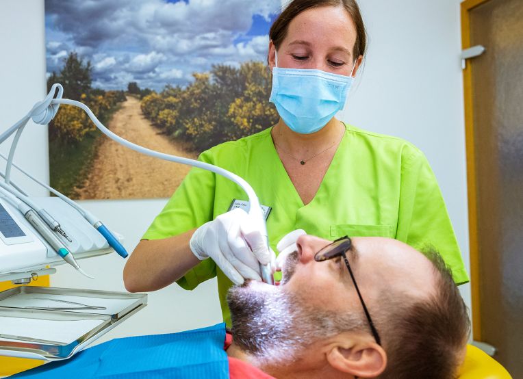 Eine Prophylaxeassistenz in grüner Kleidung und blauer Maske behandelt einen Patienten, der im Behandlungsstuhl liegt. Im Hintergrund hängt ein Landschaftsbild mit einem Weg, Büschen und bewölktem Himmel.