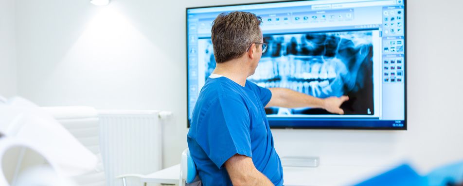 Im Fokus des Bildes steht ein Zahnarzt im blauen Kasack, der an einem großen Bildschirm auf ein Röntgenbild eines Kiefers zeigt.