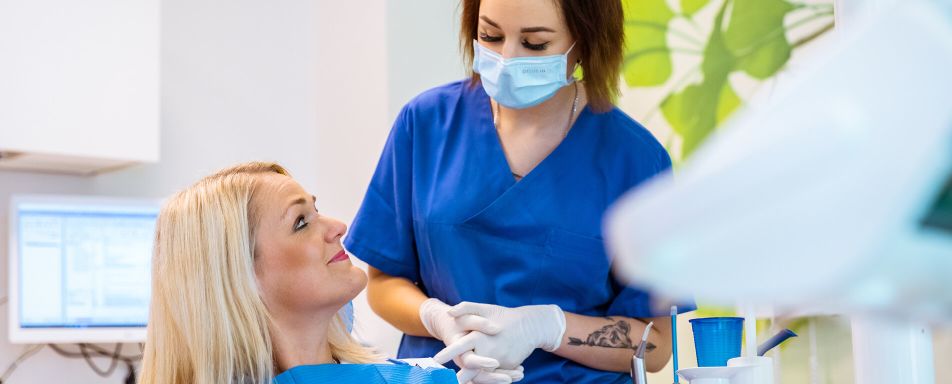 Eine Frau sitzt in einem Zahnarztstuhl, während eine weitere in blauer Kleidung, mit Maske und Handschuhen daneben steht. Im Hintergrund sind grüne Blätter als Wanddekoration zu sehen.