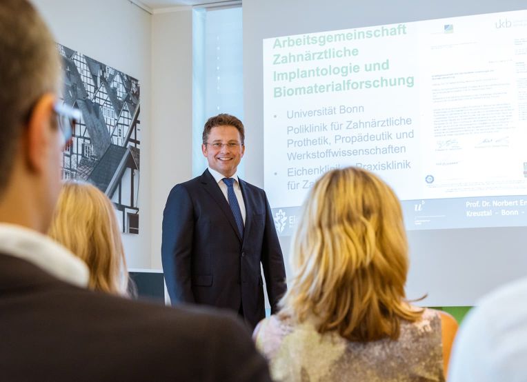 Mann in formeller Kleidung, hält einen Vortrag. Im Vordergrund sind mehrere Zuhörer zu sehen, von denen einer eine goldene Haarfarbe hat.