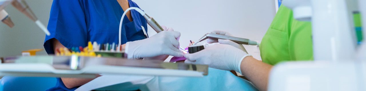 Das Bild zeigt eine zahnärztliche Behandlung in einer hellen Klinik, die von zwei Personen in grünen und blauen Kasacks mit professionellen Instrumenten durchgeführt wird.