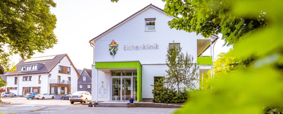Außenansicht der Eichenklinik. Das Gebäude ist weiß mit grünen Akzenten, umgeben von Bäumen und weiteren Häusern.