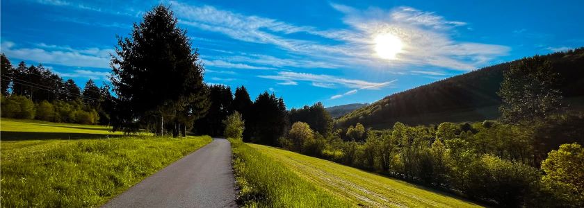 Ein geteerter Weg führt durch eine grüne Landschaft mit Bäumen, Wiesen und einem Hügel. Der Himmel ist blau und von Wolken durchzogen. Die Sonne scheint.