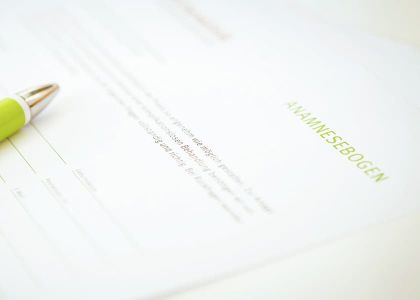 Ein Dokument mit der Überschrift „ANAMNESEBOGEN“ in grüner Schrift liegt auf einer weißen Oberfläche. Auf dem Dokument befindet sich ein grüner Kugelschreiber.