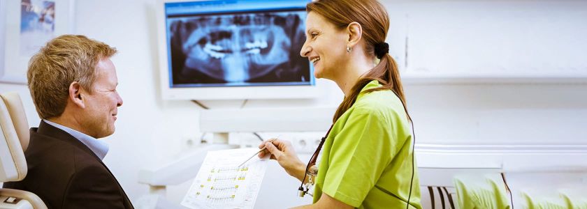 Eine zahnärztliche Fachkraft zeigt einem Patienten ein Dokument. Im Hintergrund ist ein Röntgenbild von Zähnen auf einem Monitor zu sehen.