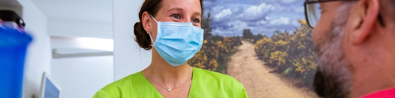 Frau in grünen Kasack mit braunen Haaren trägt einen Mund-Nasen-Schutz und spricht mit einem bärtigen Mann, der am rechten Bildrand zu sehen ist.