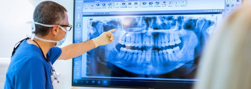 Zahnarzt zeigt auf ein großes Röntgenbild, das ein vollständiges Gebiss und den Kieferbereich darstellt. Im Vordergrund ist eine weitere Person teilweise sichtbar.