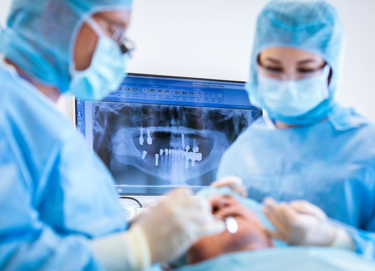 Zwei Personen in OP-Kleidung behandeln einen liegenden Patienten. Im Hintergrund zeigt ein Monitor ein Röntgenbild mit mehreren Zahnimplantaten im Kiefer.