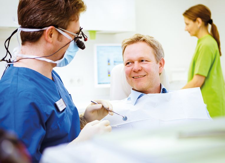 Ein Zahnarzt in blauer Berufskleidung spricht mit einem Patienten im Behandlungsstuhl.