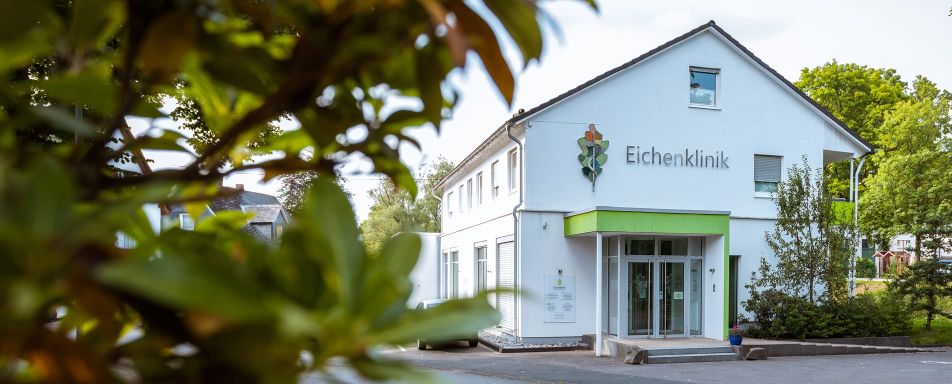 Außenansicht der Eichenklinik. Das Gebäude ist weiß mit grünen Akzenten, umgeben von Bäumen und weiteren Häusern.