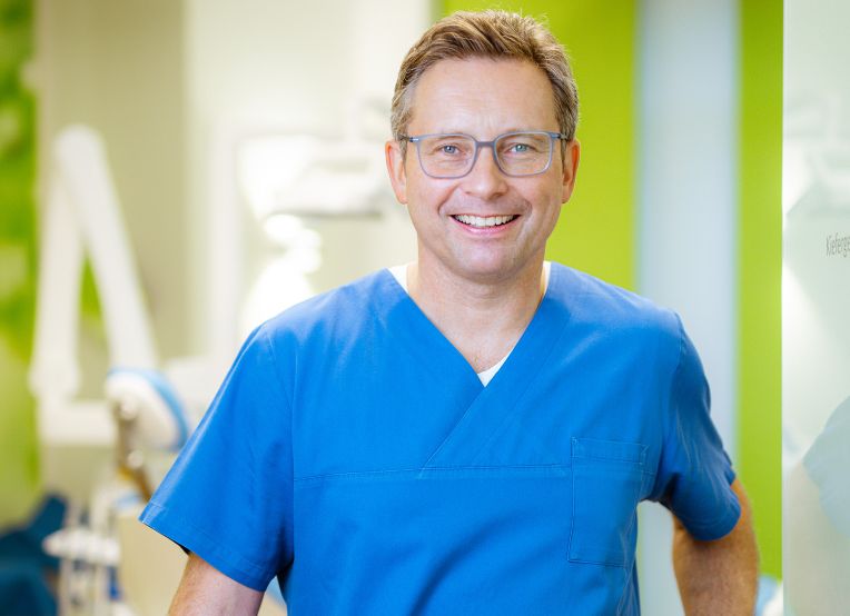 Zahnarzt im blauen Kasak, mit kurzen braunen Haaren und grauer Brille lächelt in die Kamera. Im Hintergrund ist verschwommen ein Behandlungszimmer zu erkennen.
