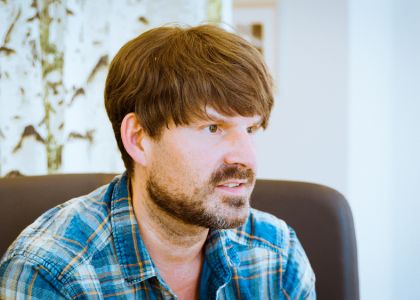 Ein Mann mit mittellangem Haar und Bart sitzt auf einem dunklen Stuhl und trägt ein blau kariertes Hemd – im Hintergrund ist eine abstrakte oder naturbezogene Wandgestaltung zu erkennen.